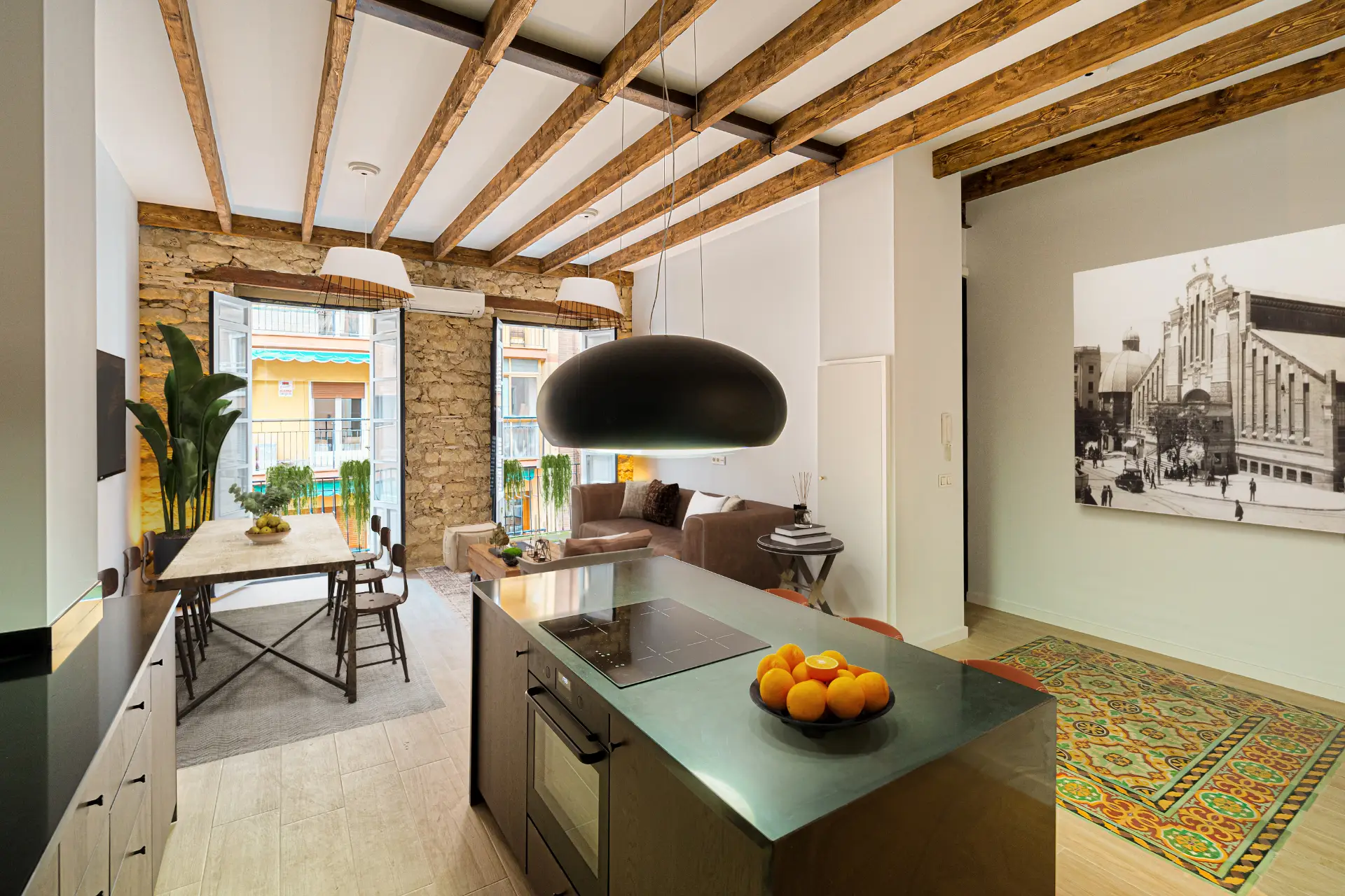 Kök och matplats med synliga träbjälkar och tegelvägg. Funktioner inkluderar en lång köksö, en stor taklampa och ett inramat fotografi på väggen.